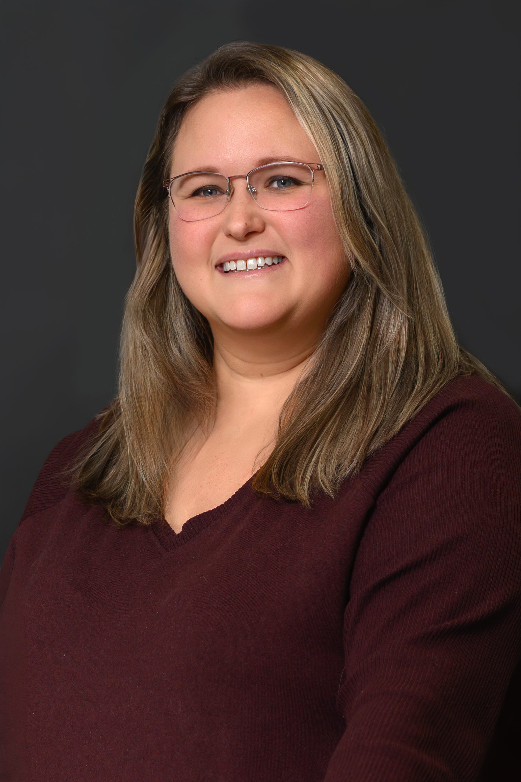 Alyssa-2026 Alyssa - A woman in front of a gray background with shoulder-length blond hair, metal frame glasses, and a dark red top.