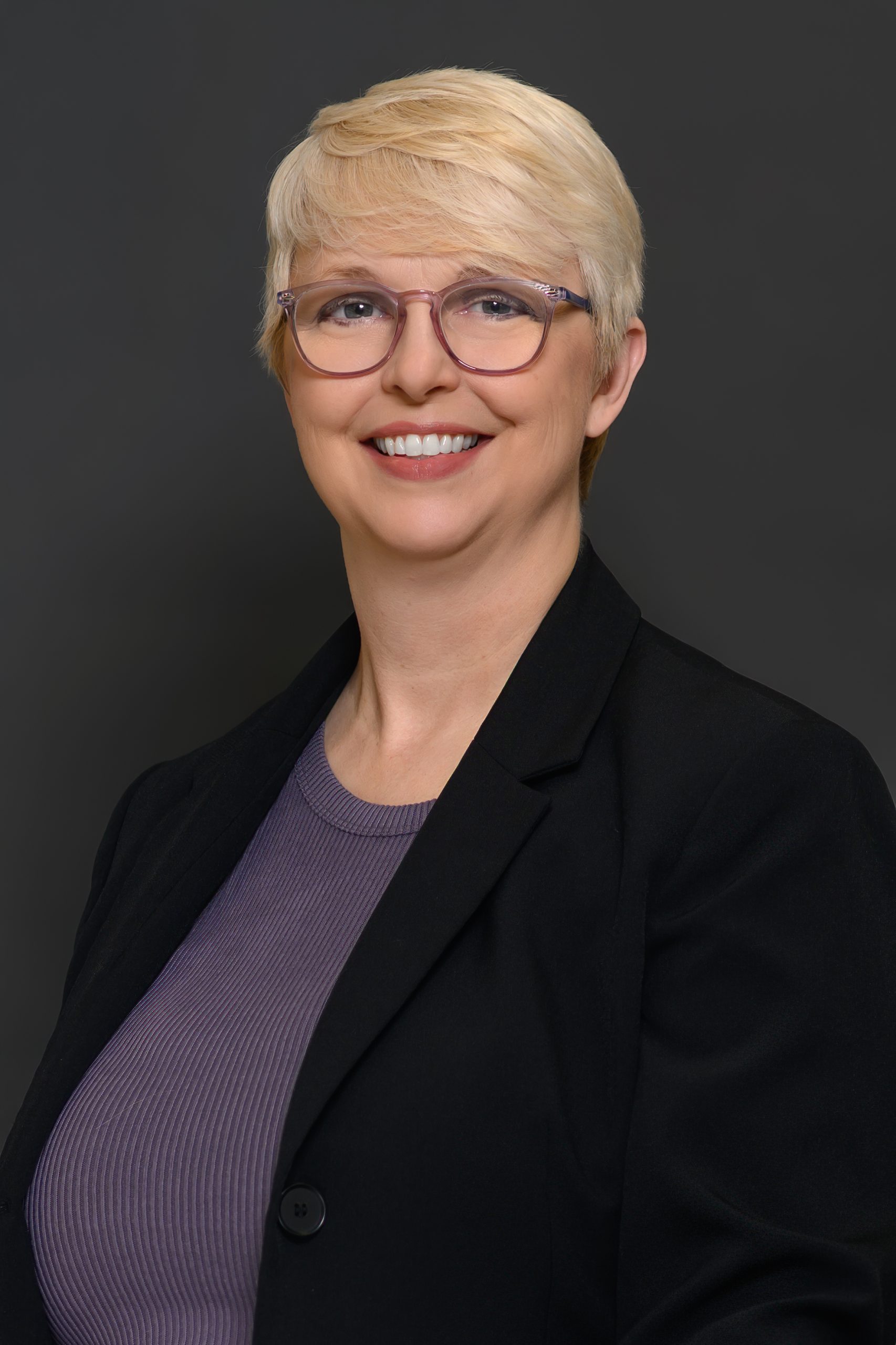 Anna-2026 Anna - A woman in front of a gray background with short blond hair, purple glasses, a purple top, and a black blazer.