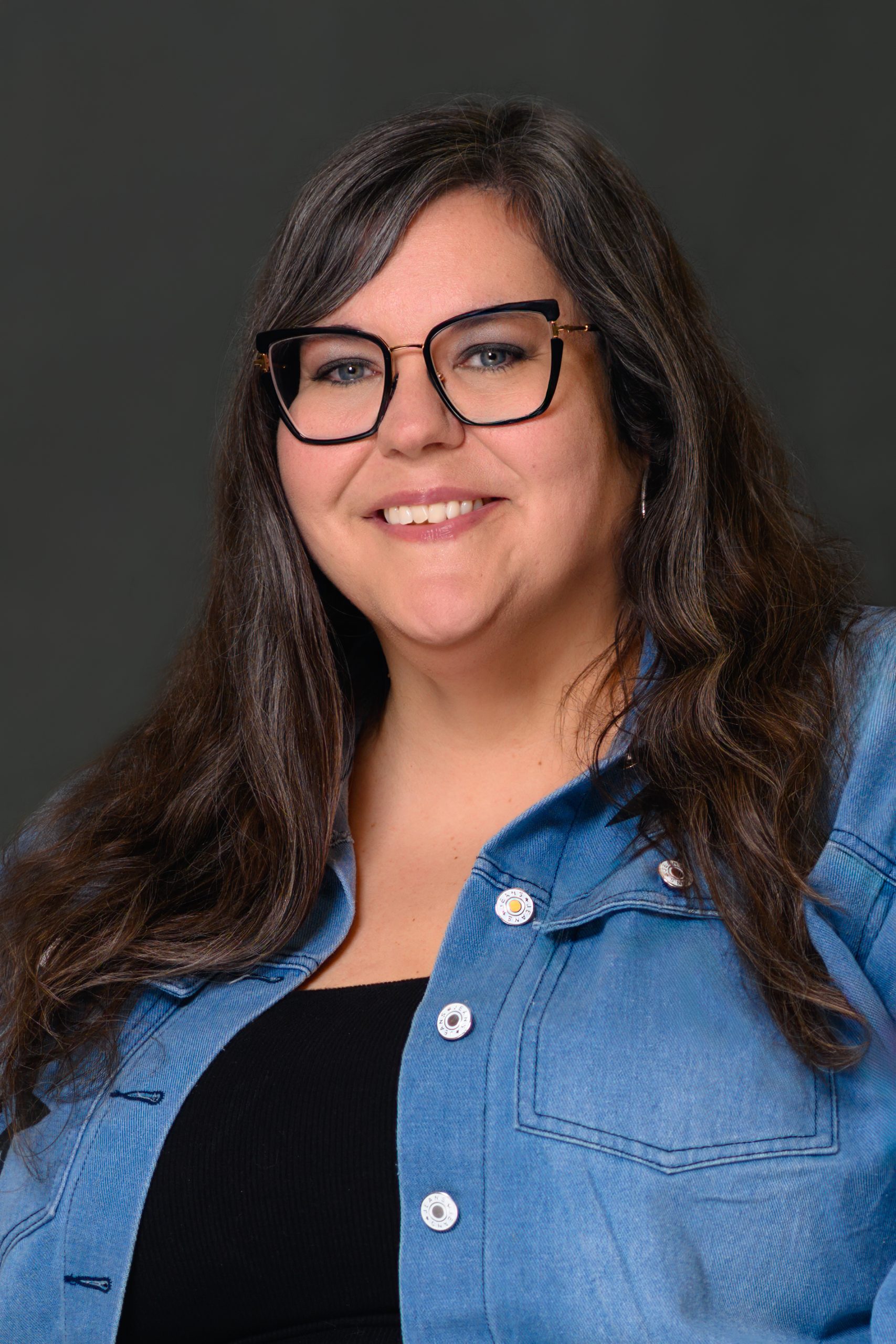 Carly-2026 Carly - A woman in front of a gray background with long dark brown hair streaked with gray, black glasses, a black top, and a denim overshirt.