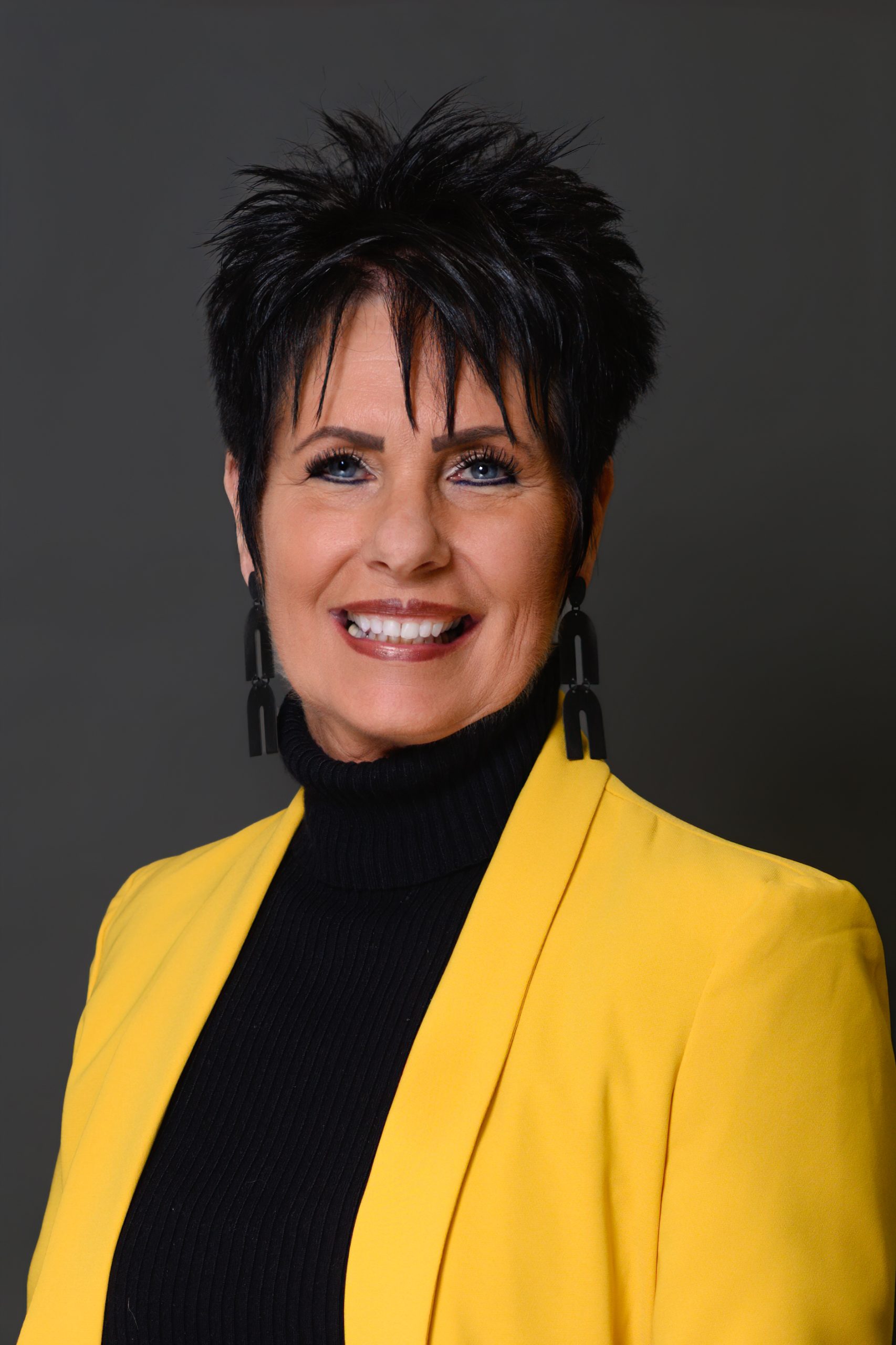 Jackie-2026 Jackie - A woman in front of a gray background with short spiky black hair, large black earrings, a black top, and a yellow blazer.